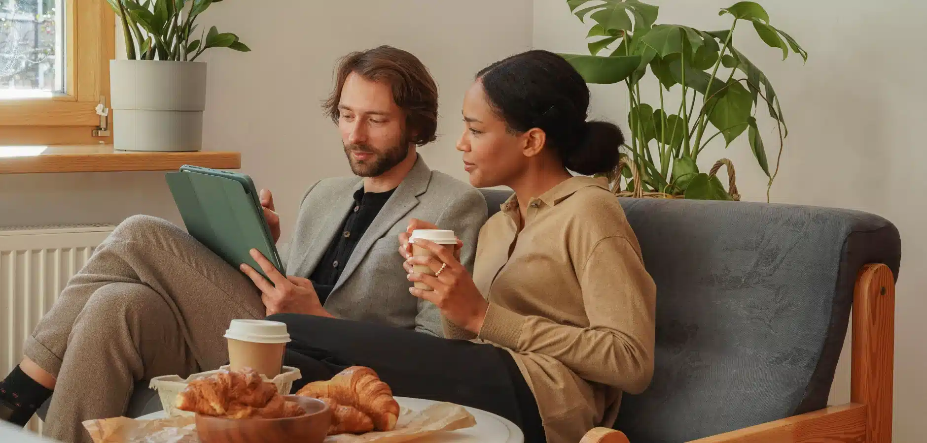 A cozy indoor scene featuring two people seated on a couch, enjoying coffee and pastries while one uses a tablet. Green plants are visible in the background.
