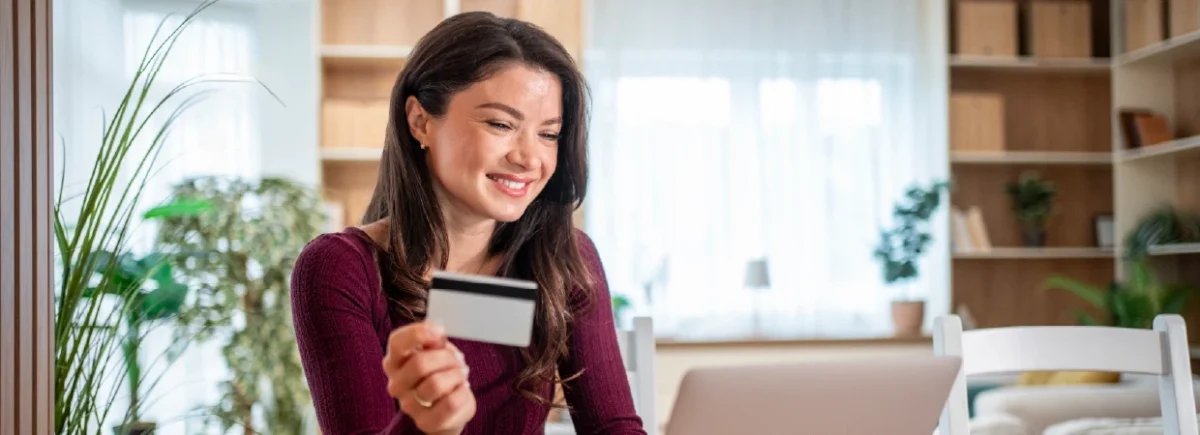 Woman holding credit card while using laptop at home with plants and shelves in the background.