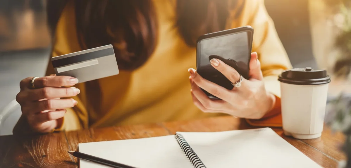 Person holding a credit card and smartphone, sitting at a table with a notebook, pen, and takeaway coffee cup.
