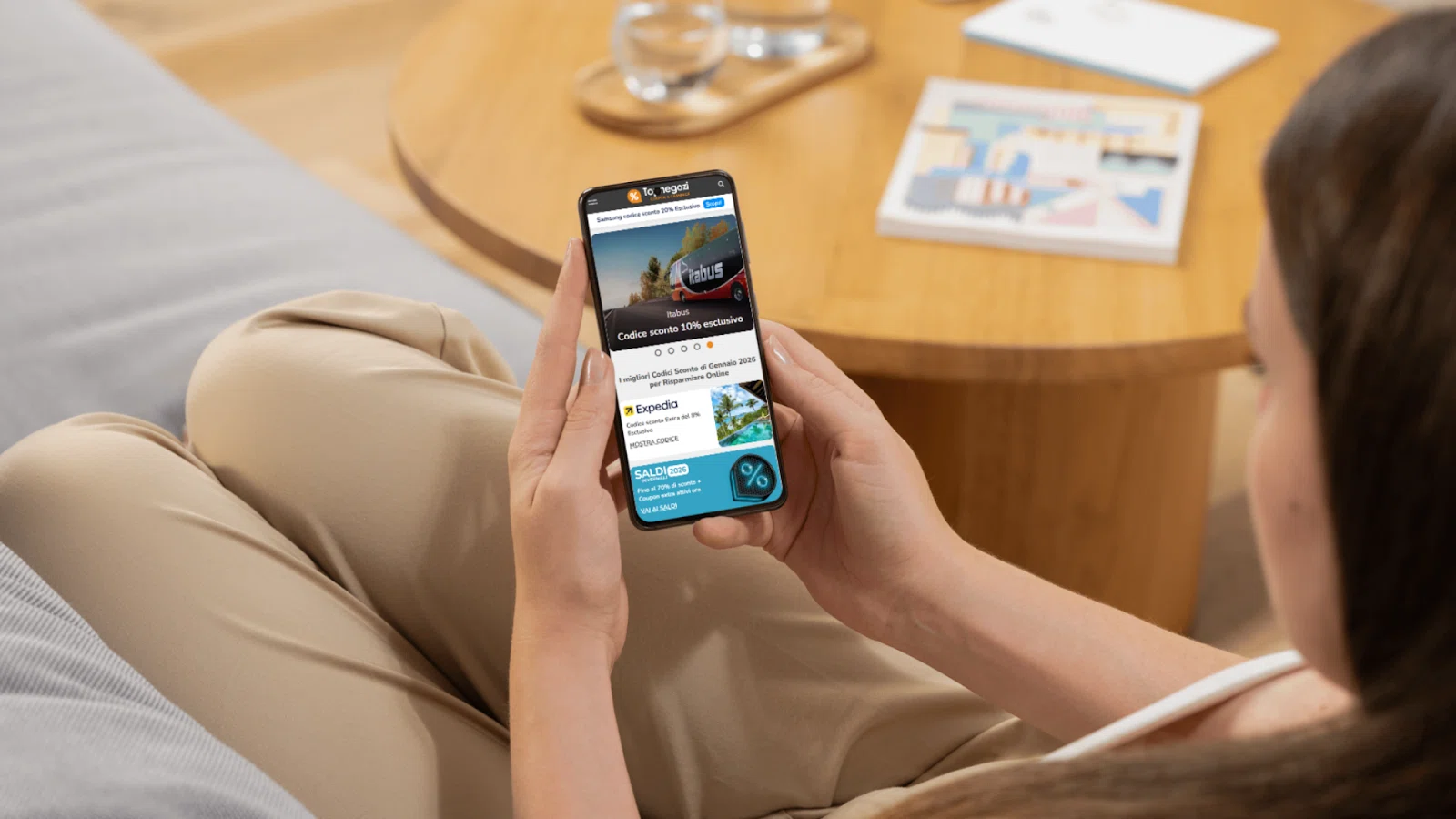 Person holding a smartphone displaying discount codes and promotional offers while sitting on a couch near a wooden table with a glass of water and a book.