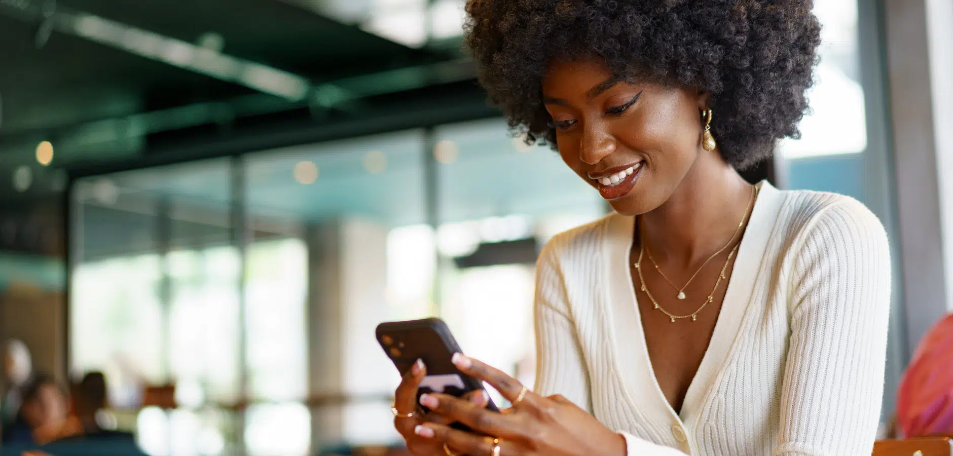 A person with curly hair sits at a table, holding a smartphone and wearing layered necklaces, in a modern indoor space.