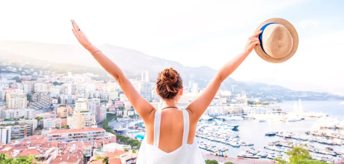 Person with arms raised, holding a hat, overlooking a coastal cityscape with boats and mountains in the background.
