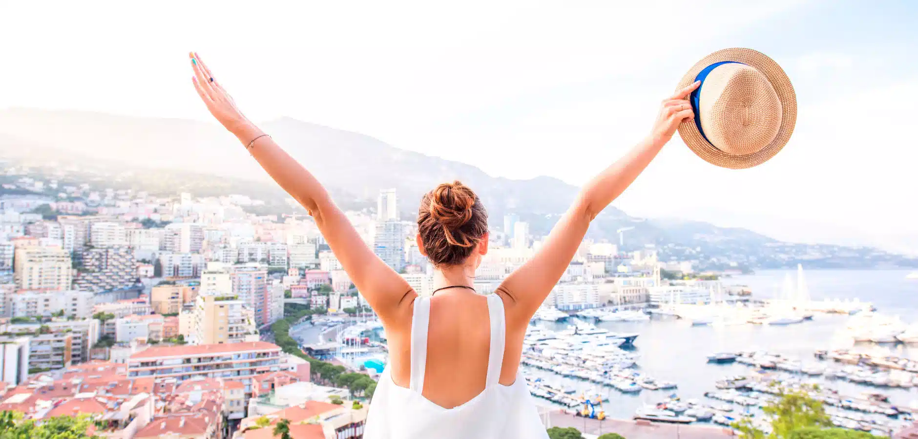 Person with arms raised, holding a hat, overlooking a coastal cityscape with boats and mountains in the background.