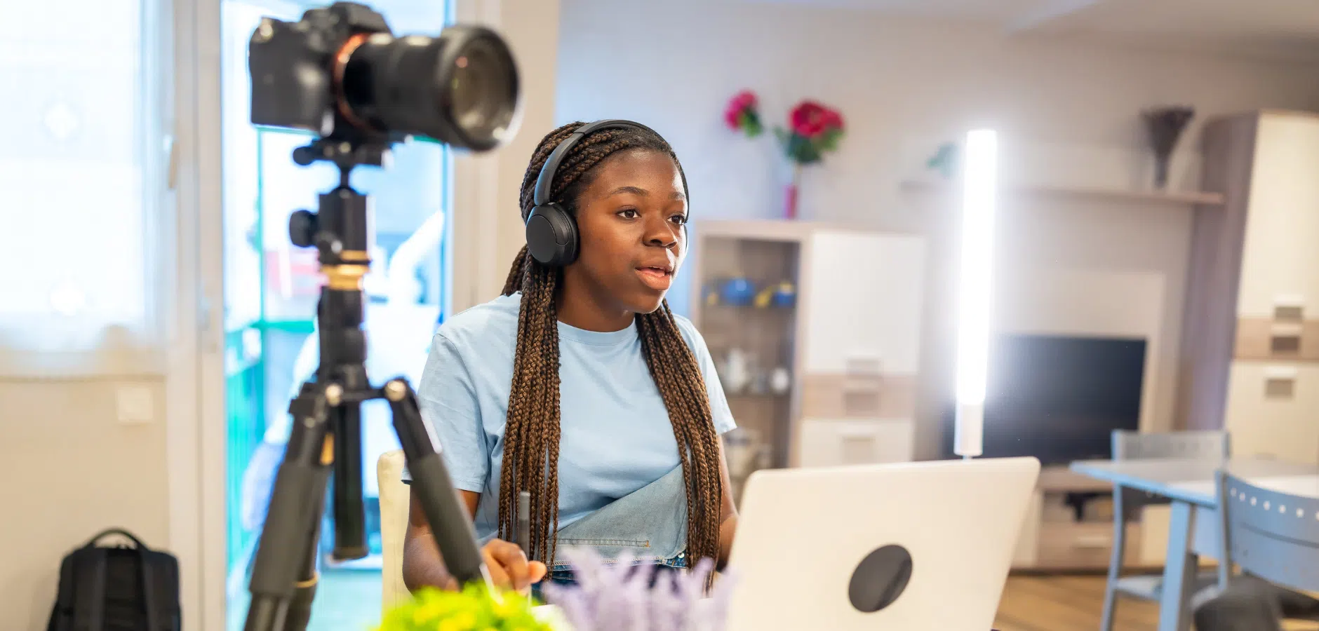 Person with long braids wearing headphones, sitting at a desk with a laptop and camera on a tripod in a bright room.