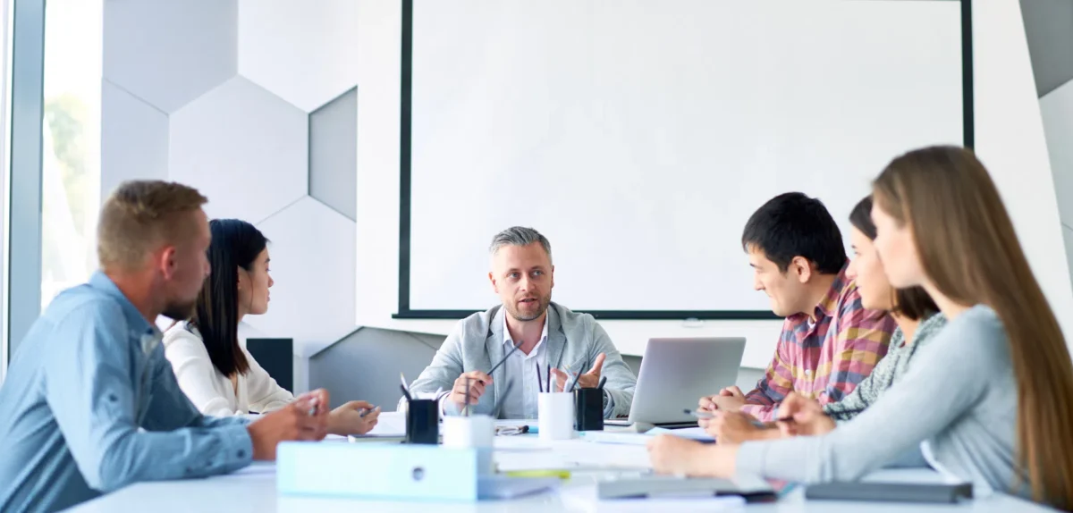 Group of six people in a meeting room, seated around a table with laptops and documents, engaged in discussion.