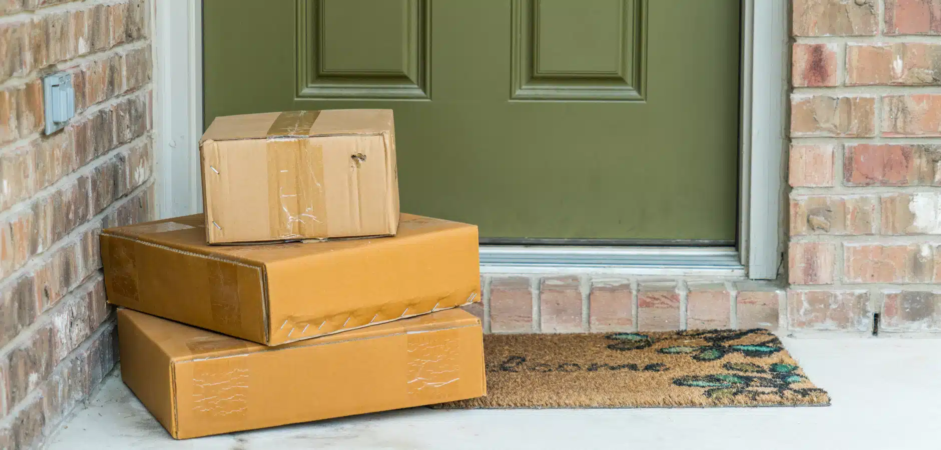 Three stacked cardboard boxes placed on a doormat in front of a green door with brick walls on either side.