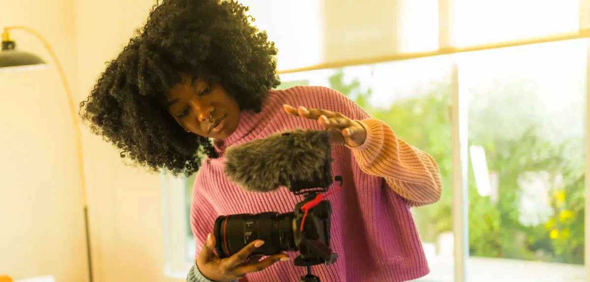 Person in a pink sweater adjusting a video camera with a furry microphone indoors near a window with green plants outside.