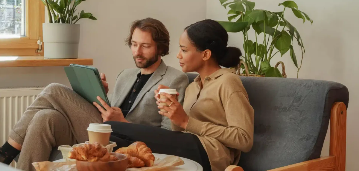 Two people sitting on a couch, one holding a tablet, the other a cup, with croissants and coffee on a table nearby and plants in the background.
