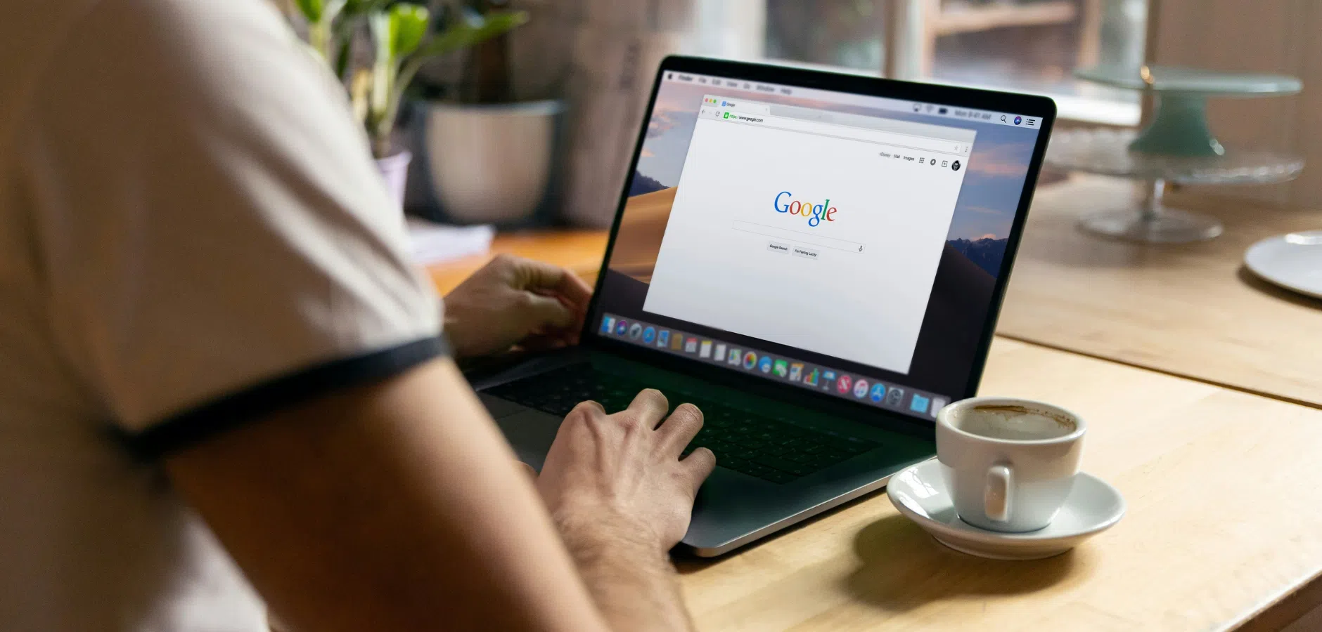 Person using a laptop with Google search open, sitting at a wooden table with a cup of coffee nearby.