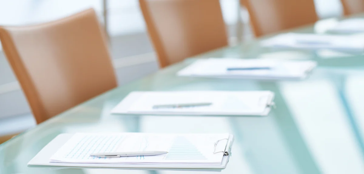 Glass conference table with clipboards, papers, and pens placed near light brown chairs in a bright office setting.