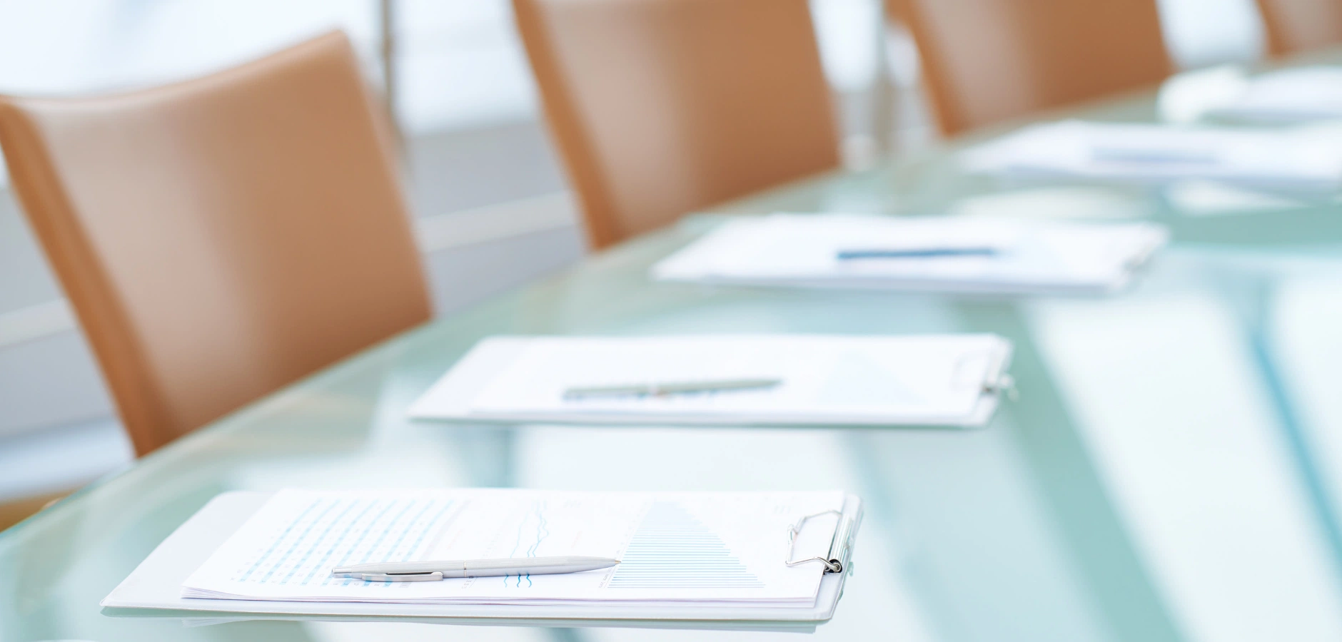Glass conference table with clipboards, papers, and pens placed near light brown chairs in a bright office setting.