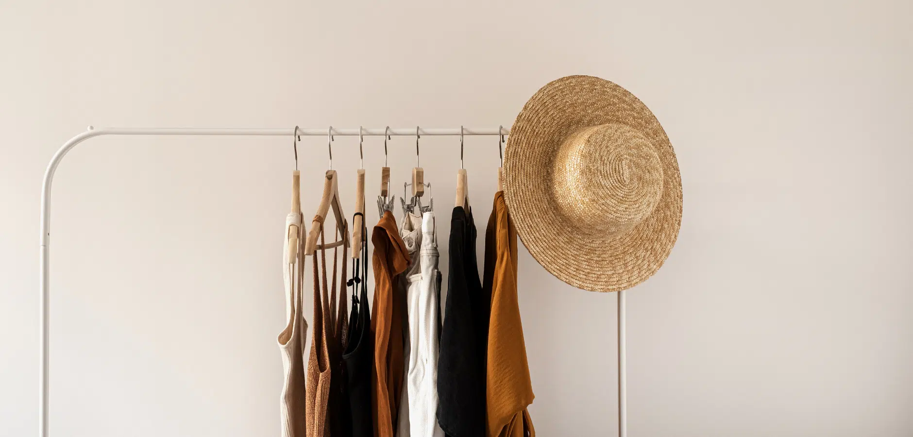 Clothing rack with neutral-toned tank tops, pants, and a large straw sunhat hanging on a white wall background.