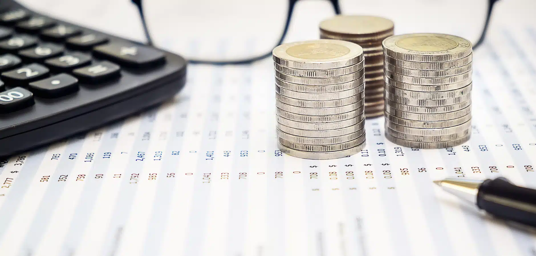 Stacks of coins, a calculator, and a pen on financial data sheets, symbolizing budgeting and accounting.