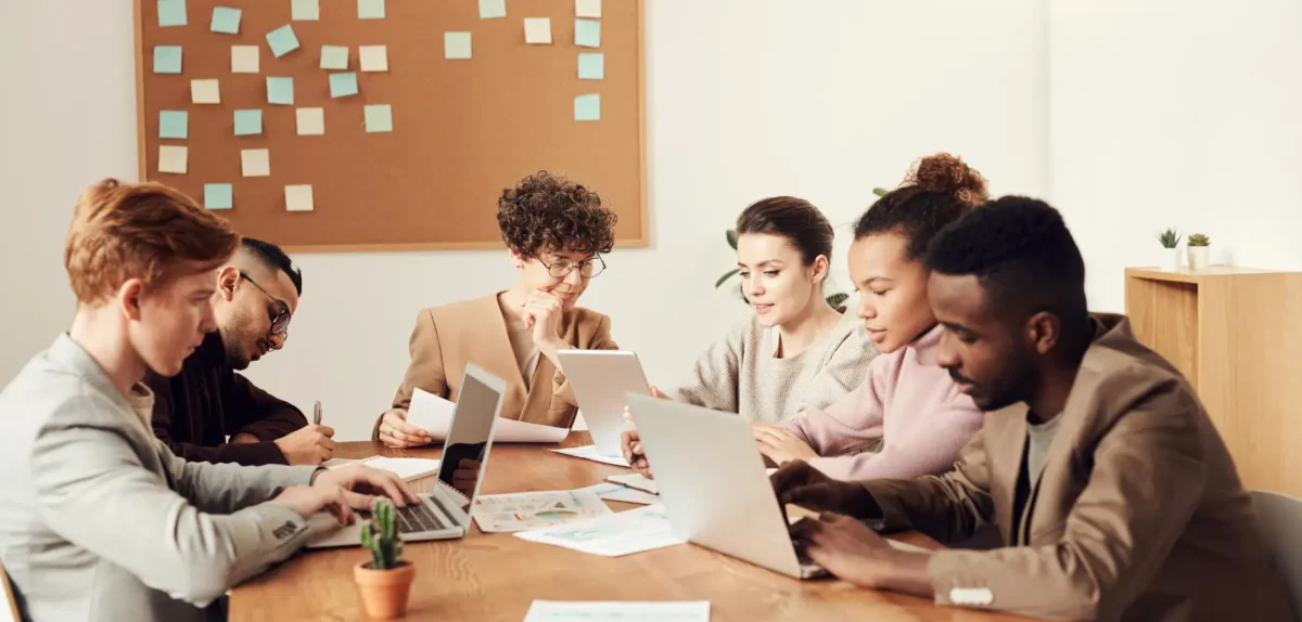 Group of six people working on laptops and tablets around a wooden table in a modern office with a corkboard and sticky notes.