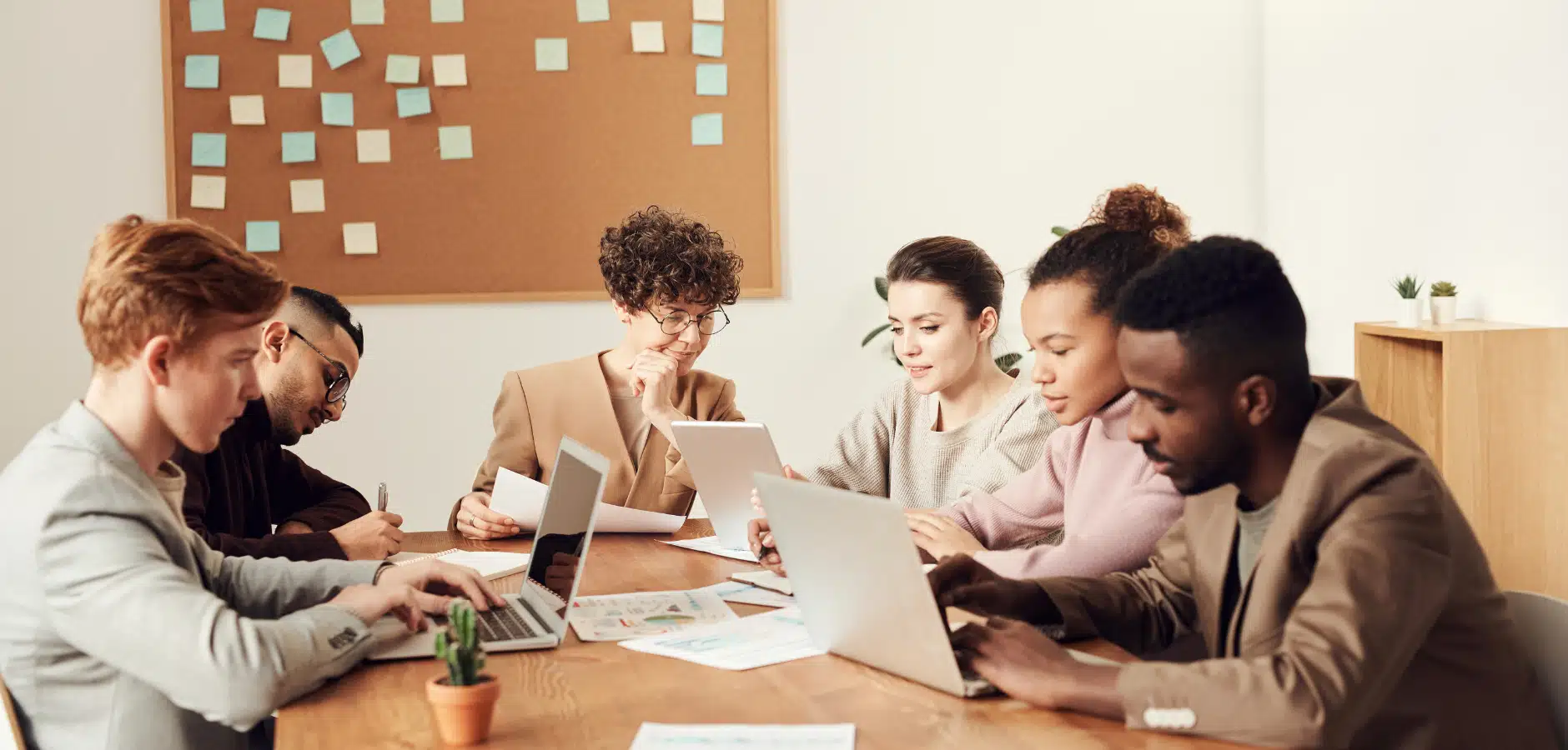 Group of six people working on laptops and tablets around a wooden table in a modern office with a corkboard and sticky notes.