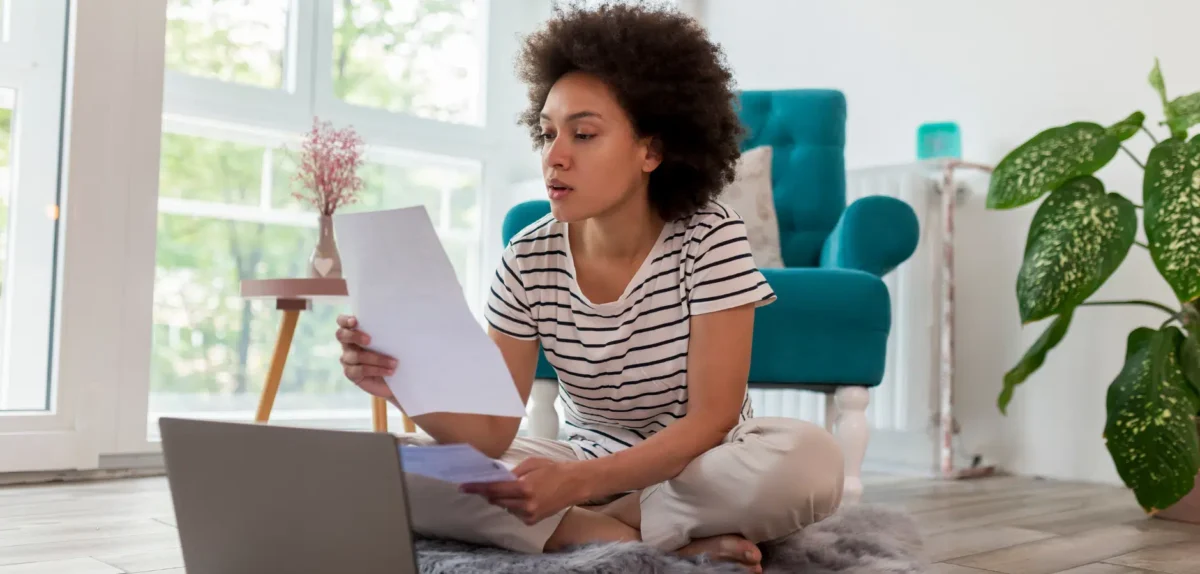 A woman seated on the floor, working on a laptop surrounded by scattered papers.