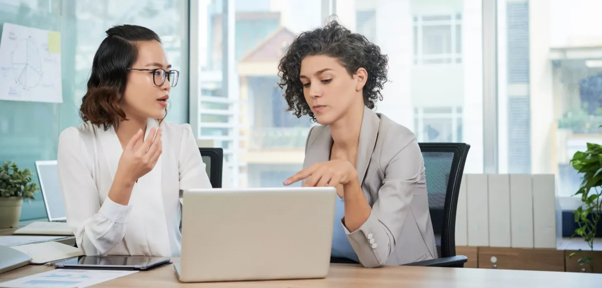 Two women collaborate at a desk, focused on a laptop, engaged in discussion and sharing ideas.