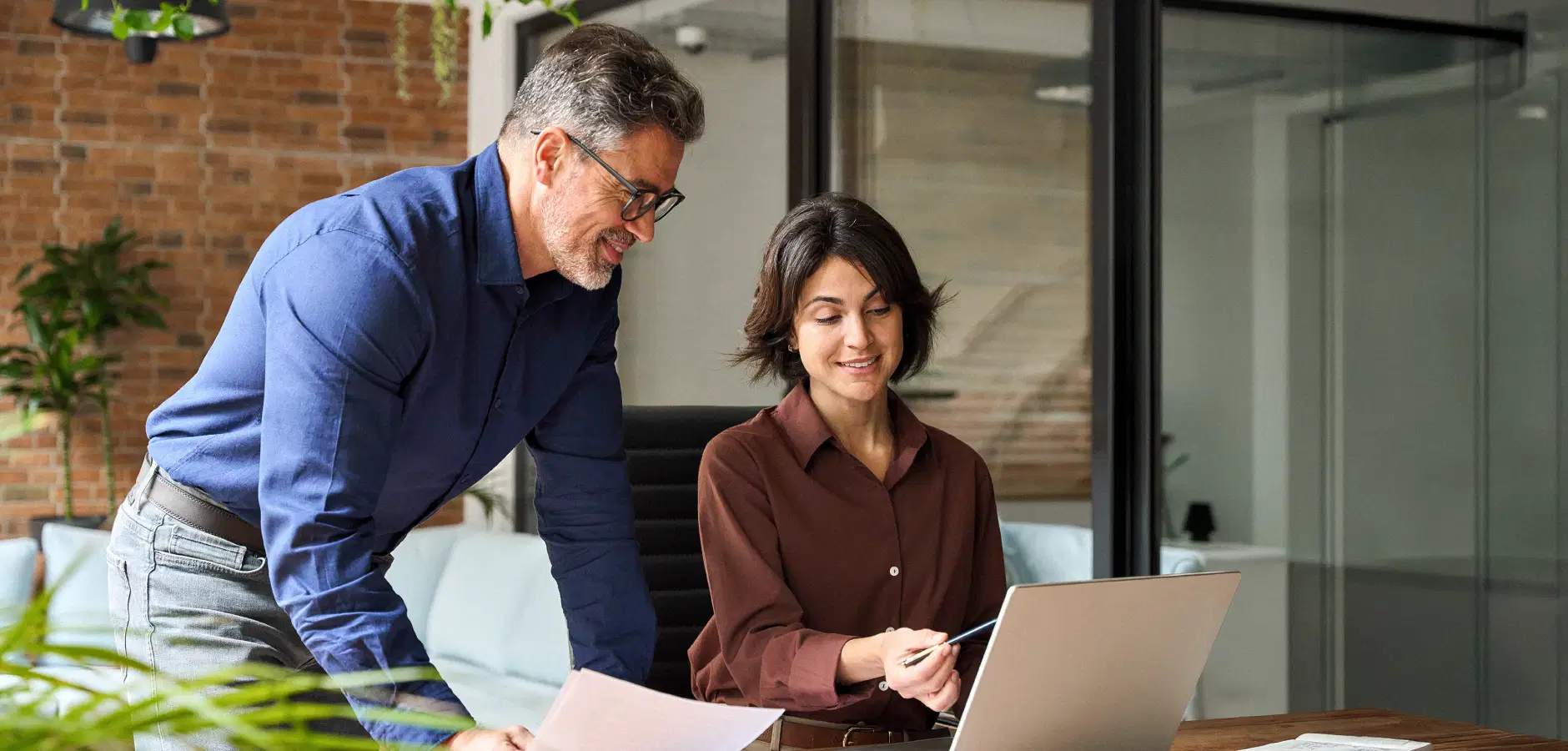 A man and woman collaborate on a laptop in a modern office setting, focused on their work.