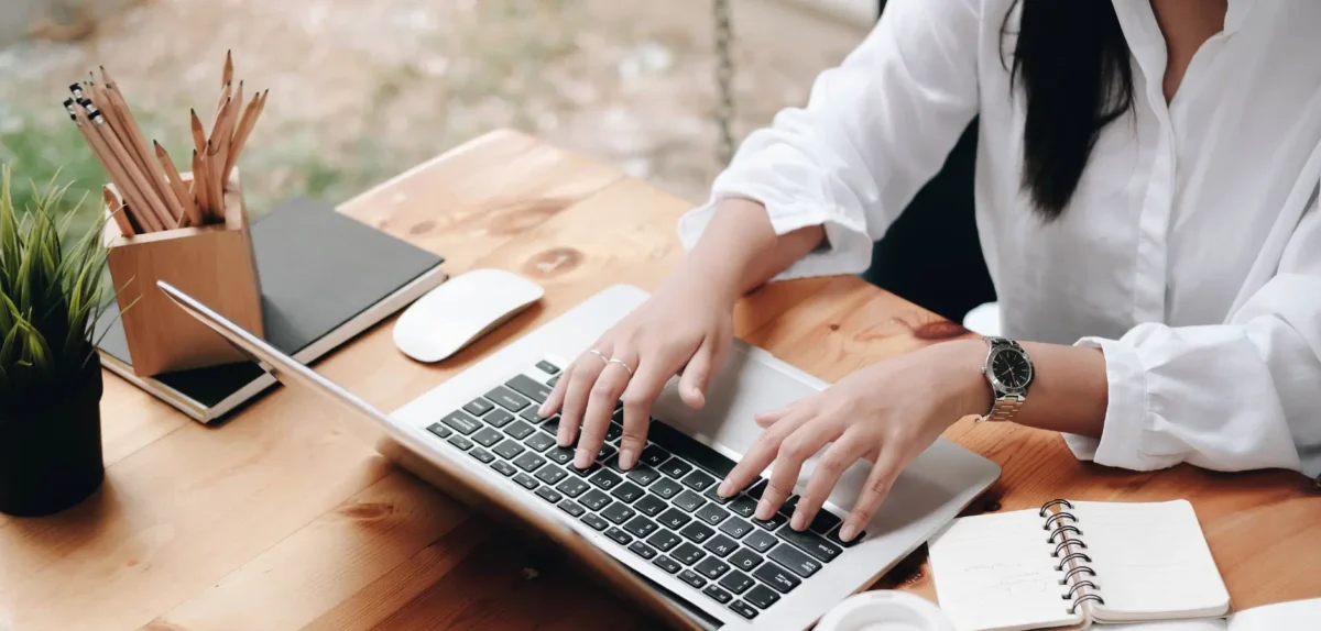 A woman focused on typing on her laptop while seated at a desk, surrounded by books and a plant.