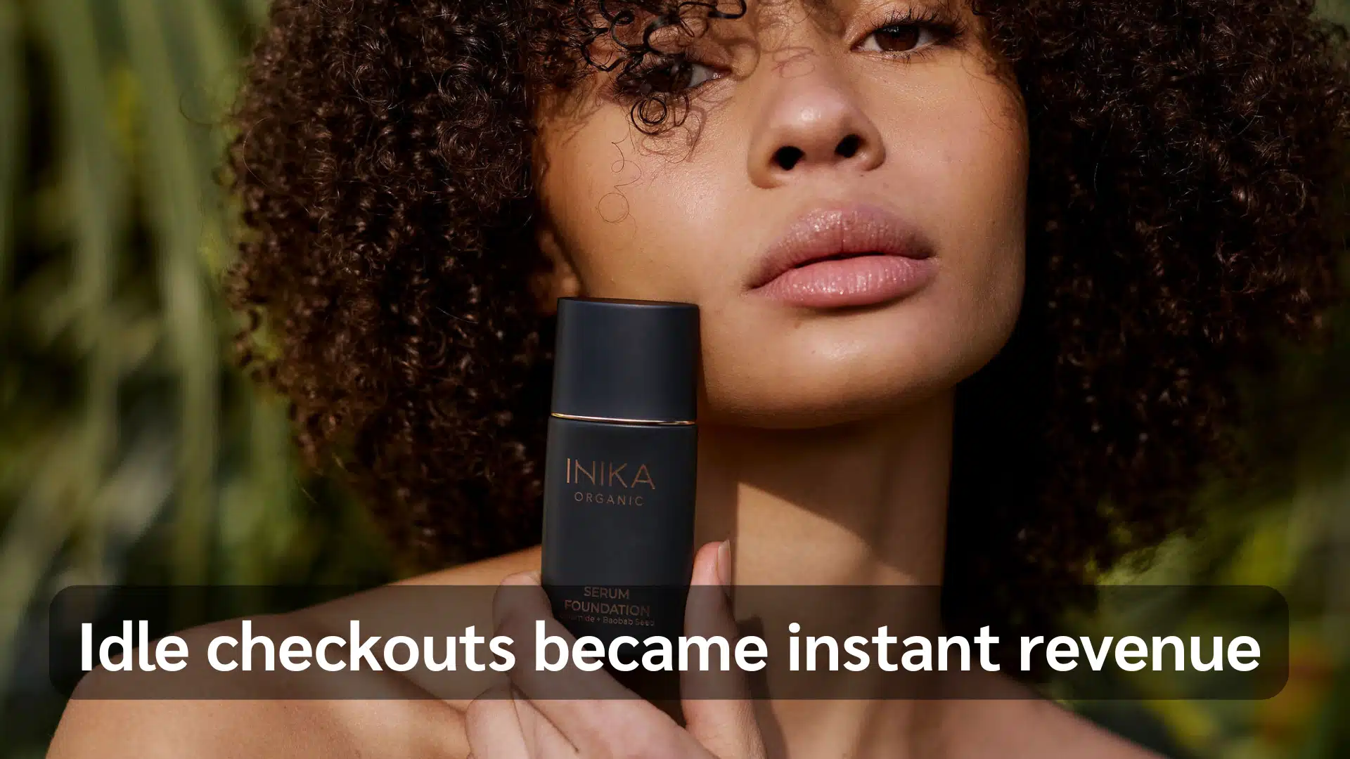 A woman with curly hair holds a bottle of skin care product, smiling and showcasing her healthy skin.
