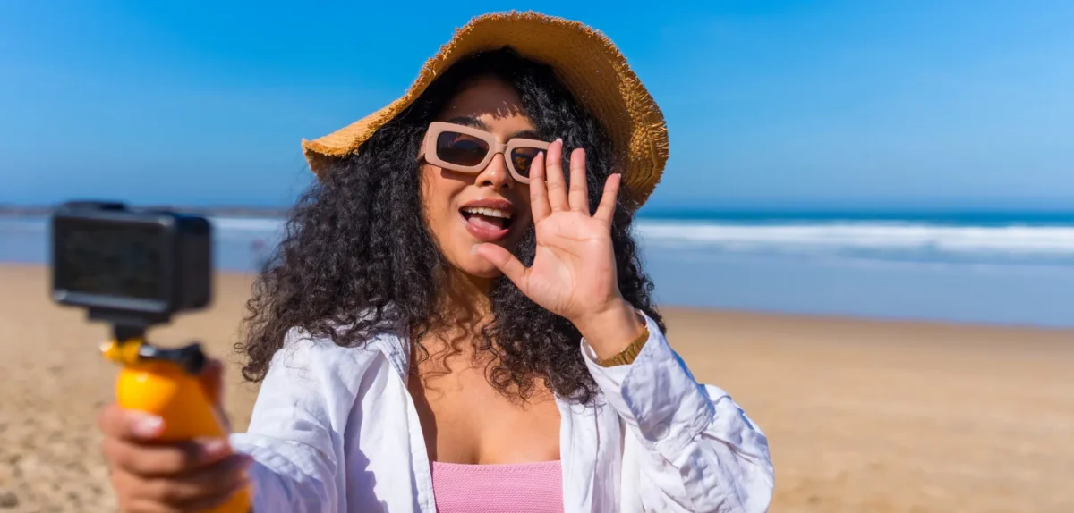 A woman wearing sunglasses and a hat takes a selfie on the beach, with the ocean and sand in the background.