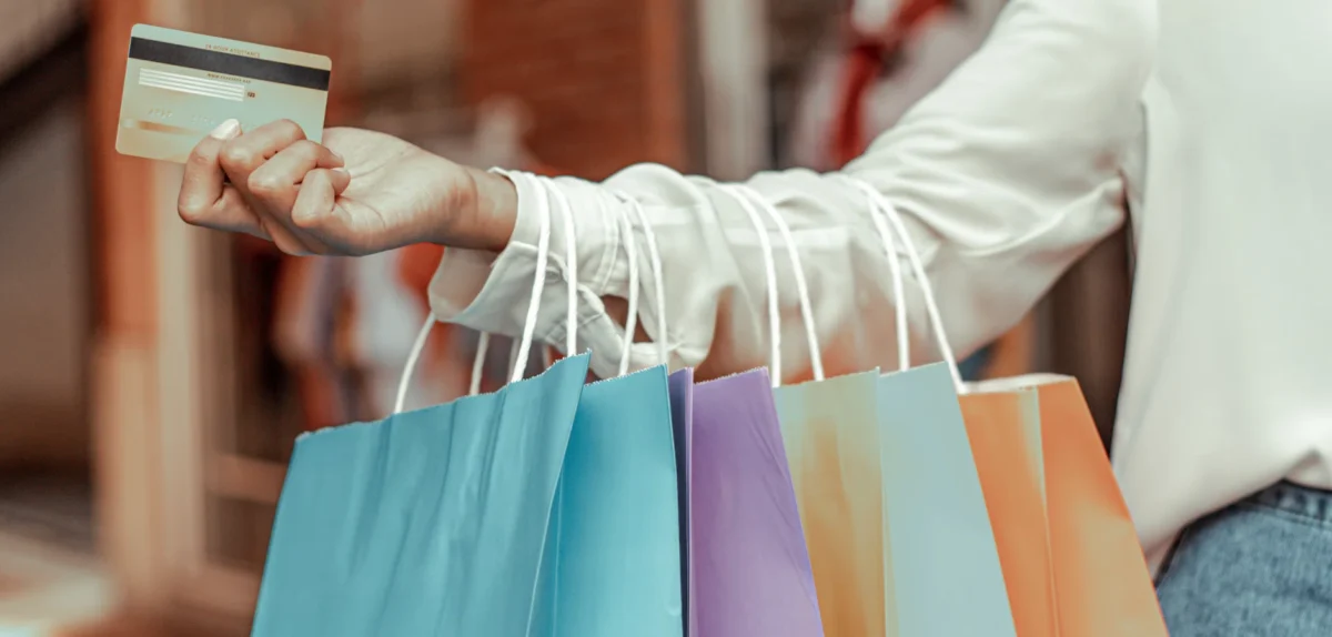 A woman holds a credit card in one hand and shopping bags in the other, smiling while shopping.