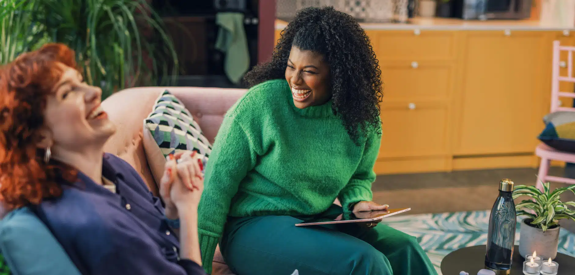 Two women laugh together in a cozy living room, with one holding a tablet and a water bottle and plants nearby.